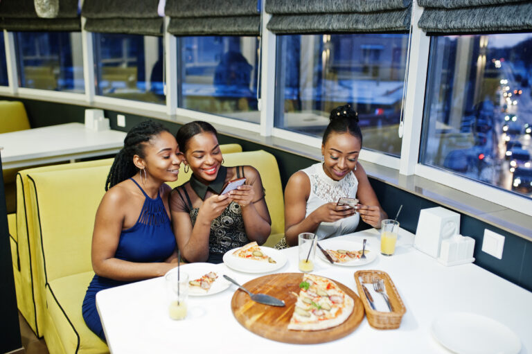 Three african woman in dress posing at restaurant, eating pizza and drink juice, making photos on phone.