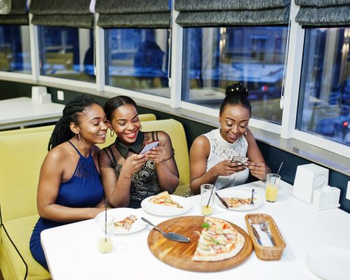 Three african woman in dress posing at restaurant, eating pizza and drink juice, making photos on phone.